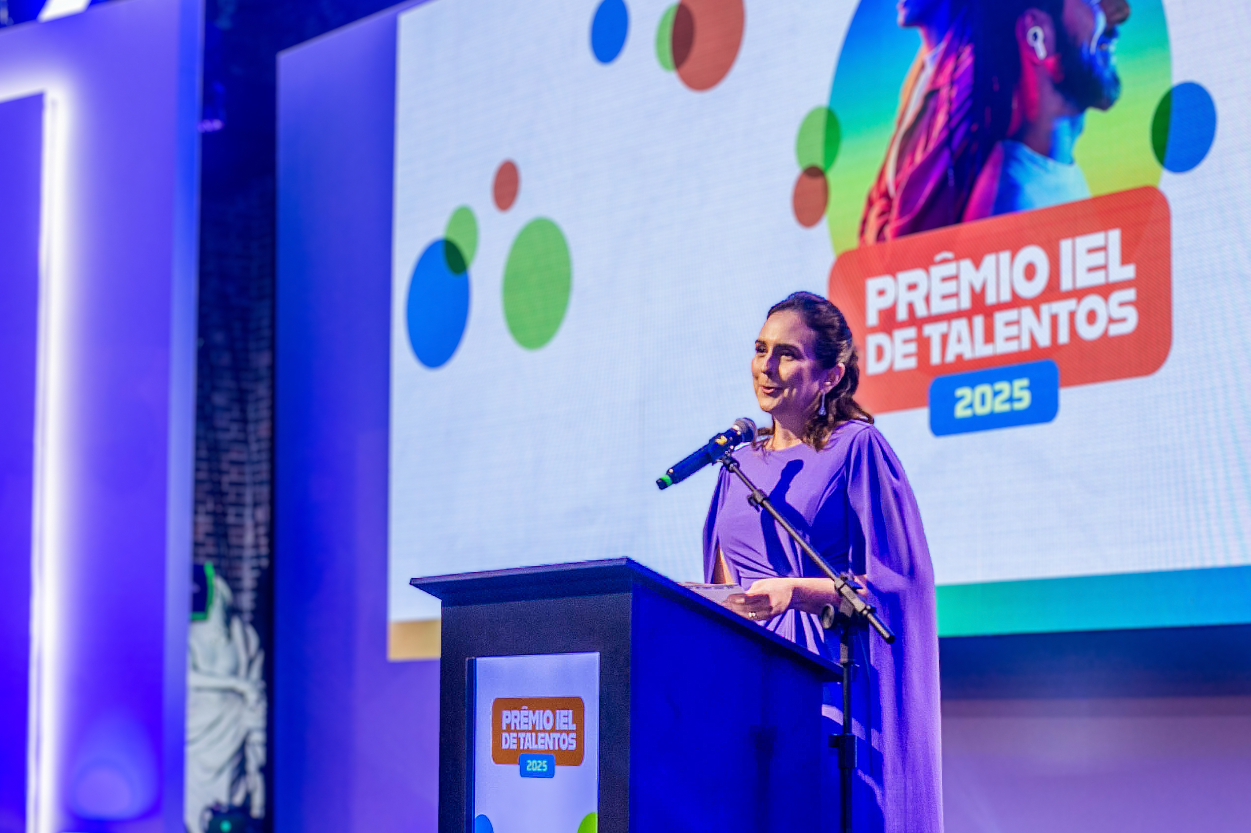 Uma mulher sorridente com cabelos longos e escuros está em pé em uma sala de escritório moderna e falando com entusiasmo, gesticulando com as mãos. Ela veste uma blusa vermelha e calças escuras. Duas outras mulheres estão sentadas à mesa. Uma delas tem cabelos loiros e veste um blazer bege, enquanto a outra tem cabelos crespos e escuros e veste um blazer rosa. O ambiente sugere uma apresentação ou reunião de negócios.