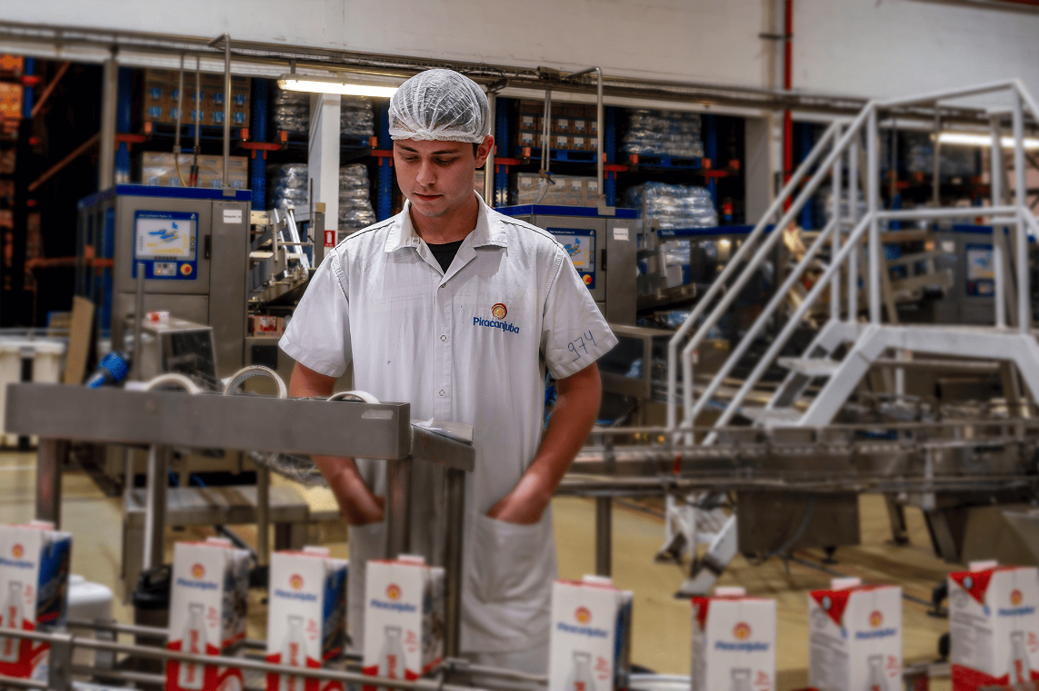 Em um ambiente industrial, um jovem com pele clara e gorro branco inspeciona embalagens de papelão que se movem em uma esteira rolante. Ele veste um uniforme branco.
